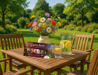 Holztisch im Garten mit buntem Blumenstrauß in einer Vase, daneben eine Karaffe und ein Glas mit Zitronenwasser sowie ein Gutschein im Vordergrund.