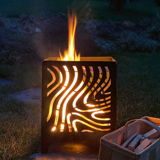 Feuerkorb mit dekorativem Wellenmuster, leuchtendem Feuer und Holzvorratskiste daneben, aufgenommen bei Nacht im Freien.