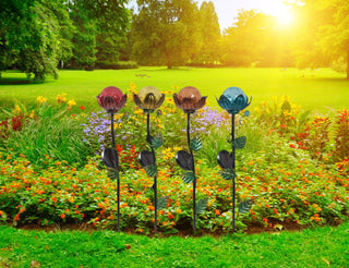Garten mit grünem Rasen und bunten Blumen im Hintergrund. Vier Metall-Gartenstecker mit gläsernen Blüten in Rot, Gelb, Orange und Blau stehen in einer Reihe und setzen farbige Akzente vor dem natürlichen Grün.