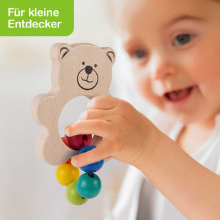 Ein Kind hält eine Holzrassel in Form eines lächelnden Teddybären. Die Rassel hat eine runde Öffnung in der Mitte und ist mit bunten Kugeln in Rot, Blau, Grün und Gelb verziert. In der oberen linken Ecke steht der Schriftzug „Für kleine Entdecker“.