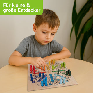 Kind spielt konzentriert ein buntes Brettspiel mit Spielfiguren am Tisch. Oben links steht: ‚Für kleine & große Entdecker‘ auf grünem Hintergrund. Im Hintergrund ist eine Zimmerpflanze sichtbar.