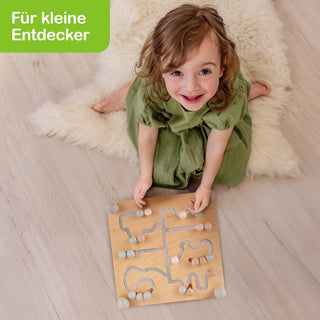 Kleines Kind spielt auf flauschigem Teppich mit einem bunten Holzlabyrinth-Spiel. Schriftzug ‚Für kleine Entdecker‘ oben links auf grünem Hintergrund. Szene vermittelt Spielspaß und frühe Förderung.
