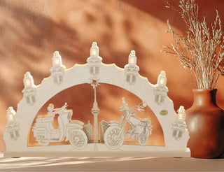 Holzbogen mit filigranen Ausschnitten von zwei Motorrädern und einer Stadtsilhouette mit Turm, beleuchtet von sieben kleinen Lampen. Im Hintergrund warme Wand, rechts eine Vase mit Trockenpflanzen.