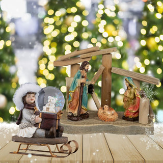 Weihnachtliche Szene mit Krippe, Figuren von Maria, Josef und Jesuskind unter einem Holzstall, im Vordergrund eine Figur mit Schlitten und Schneekugel, im Hintergrund verschwommene Weihnachtslichter und dekorierte Tannen.