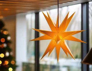 Eine leuchtende, gelbe sternförmige Laterne (Adventsstern) hängt von der Decke in einem modernen, gemütlichen Raum mit großen Fenstern. Im Hintergrund ist ein verschwommener Blick auf einen geschmückten Weihnachtsbaum mit Lichtern und Ornamenten zu sehen.