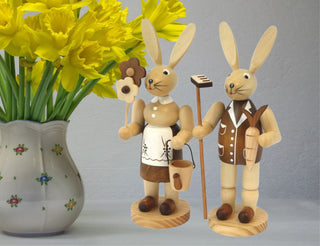 Zwei Holzosterhasen in Tracht stehen neben gelben Narzissen. Sie halten Blume, Eimer, Karotte und Rechen. Die Szene zeigt Frühlingsstimmung mit Sonnenschein.