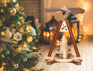 Holz-Weihnachtspyramide mit zwei brennenden Kerzen, kleinen Tannenbäumen und einem Weihnachtsmann in der Mitte. Im Hintergrund steht ein verschwommener, beleuchteter Weihnachtsbaum.