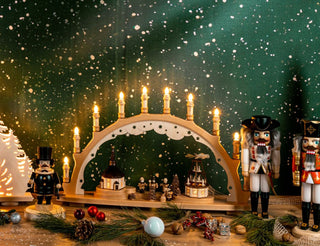 Weihnachtliche Szene mit beleuchtetem Schwibbogen, Nussknackern, Miniaturhäusern, Tannenzweigen, Kugeln und Zapfen vor grünem Hintergrund mit Schneeflocken auf einem Holztisch.