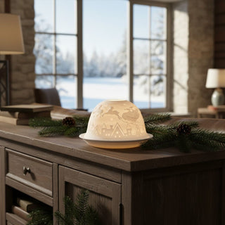 Kerzenhalter „Dome Light“ aus weißem Porzellan für ein Teelicht, auf einem Holztisch dekoriert. Die halbrunde Kuppel zeigt ein filigranes Motiv mit einer winterlichen Stadt und einer Schlittenfahrt mit Pferd darüber. Durch das warme Kerzenlicht leuchten die feinen Konturen sanft auf. Im Hintergrund ist ein Fenster mit verschneiter Landschaft zu sehen; auf dem Tisch liegen Tannenzweige und Zapfen als winterliche Dekoration.