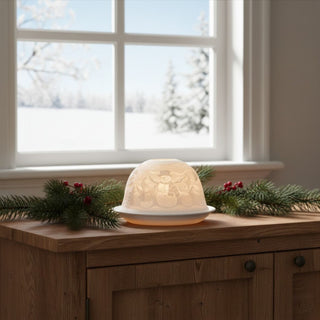 Kerzenhalter „Dome Light“ aus weißem Porzellan für ein Teelicht, auf einem Holzmöbel dekoriert. Die halbrunde Kuppel zeigt ein filigranes Schneemannmotiv mit Hut und Schal, umgeben von winterlichen Elementen. Durch das warme Kerzenlicht werden die feinen Details sanft beleuchtet. Im Hintergrund ist ein Fenster mit verschneiter Landschaft zu sehen; auf dem Möbel liegen Tannenzweige mit roten Beeren als festliche Dekoration.