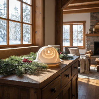 Kerzenhalter „Dome Light“ aus weißem Porzellan für ein Teelicht, auf einer Holzkommode dekoriert. Die halbrunde Kuppel zeigt ein modernes Flammenmotiv mit konzentrischen, weich verlaufenden Formen, die durch das warme Kerzenlicht sanft leuchten. Im Hintergrund ist ein gemütlicher Wohnraum mit großen Fenstern und verschneiter Landschaft zu sehen; auf der Kommode liegen Tannenzweige und rote Beeren als winterliche Dekoration.