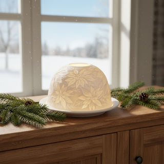 Kerzenhalter „Dome Light“ aus weißem Porzellan für ein Teelicht, auf einer Holzkommode dekoriert. Die halbrunde Kuppel zeigt ein filigranes Poinsettia‑Motiv mit Weihnachtssternen, das durch das warme Kerzenlicht sanft beleuchtet wird. Im Hintergrund ist ein Fenster mit verschneiter Landschaft zu sehen; auf der Kommode liegen Tannenzweige und Zapfen als winterliche Dekoration.