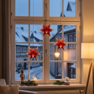 Marienberger Adventsstern 3er-Set in Rot-Gelb mit 15 cm Durchmesser. Drei kleine, mehrzackige Weihnachtssterne hängen an feinen Schnüren vor einem Fenster und leuchten in warmen Rot- und Gelbtönen. Auf der Fensterbank sorgen eine Kerze, ein Nussknacker und winterliche Dekoration für eine festliche Atmosphäre. Im Hintergrund ist eine verschneite Altstadt mit beleuchteter Straßenlaterne zu sehen.