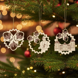 Baumbehang „Drei Haselnüsse für Aschenbrödel®“ im 3er Set, farbig abgesetzte Holzornamente mit Haselnuss-Motiv, Figuren und Schloss, am Weihnachtsbaum aufgehängt, umgeben von grünen Tannenzweigen und warm leuchtenden Lichtern.
