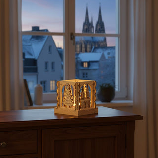 Kleiner Kerzenhalter aus naturfarbenem Holz für ein Teelicht mit Motiv „Köln“, auf einer Holzkommode platziert. Der quadratische Halter zeigt filigrane Ausschnitte mit dem Kölner Dom und einem stilisierten Weihnachtsbaum, die vom warmen Kerzenlicht im Inneren beleuchtet werden. Im Hintergrund ist ein Fenster mit verschneiter Stadtszene zu sehen, in der Ferne ragt ebenfalls der Dom auf; innen stehen Bücher und ein Sessel.