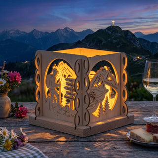 Natürlicher Holz-Kerzenhalter „Wanderfreund“ für ein Teelicht (ca. 12 cm), würfelförmig mit filigranen Motiven eines Wanderers, Wegweisers und einer alpinen Berglandschaft. Der warm leuchtende Kerzenhalter steht auf einem Holztisch im Freien bei Abenddämmerung; im Hintergrund sind Berge mit Gipfelkreuz zu sehen. Auf dem Tisch befinden sich zudem Blumen, ein Weinglas und eine Brotzeit, die eine gemütliche Bergatmosphäre schaffen.