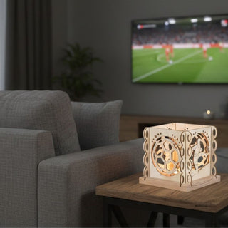 Kerzenhalter aus naturfarbenem Holz für ein Teelicht mit Motiv „Fußballfreunde“, auf einem Beistelltisch im Wohnzimmer platziert. Der quadratische Halter zeigt filigrane Ausschnitte mit Fußballmotiven wie Ball und Spielerfiguren, die vom warmen Kerzenlicht im Inneren beleuchtet werden. Im Hintergrund stehen ein Sessel und ein Fernseher mit laufendem Fußballspiel; der Raum ist modern und gemütlich eingerichtet.
