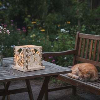 Kerzenhalter aus naturfarbenem Holz für ein Teelicht mit Motiv „Katzenfreund“, dekoriert auf einem Holztisch im Garten. Der quadratische Halter zeigt filigrane Ausschnitte mit Katzen und verspielten Ornamenten, die vom warmen Kerzenlicht im Inneren beleuchtet werden. Neben dem Tisch liegt eine ruhende Katze auf einer Holzbank; im Hintergrund sind Blumen und grüne Pflanzen in einer sommerlichen Gartenszene zu sehen.