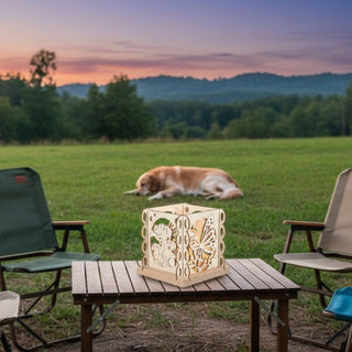 Kerzenhalter aus naturfarbenem Holz für ein Teelicht mit Motiv „Hundefreund“, auf einem kleinen Holztisch im Freien platziert. Der quadratische Halter zeigt filigrane Ausschnitte mit Hundemotiven und dekorativen Elementen, die vom warmen Kerzenlicht im Inneren beleuchtet werden. Im Hintergrund liegt ein Hund auf einer Wiese, umgeben von einer weiten Landschaft mit Bäumen und Hügeln im Abendlicht; daneben stehen zwei Gartenstühle.