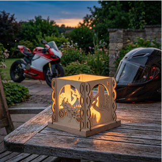 Natürlicher Holz-Kerzenhalter „Motorradfreund“ für ein Teelicht (ca. 12 cm), würfelförmig mit filigranen Motorrad- und Landschaftsmotiven. Der leuchtende Kerzenhalter steht auf einem Holztisch im Garten bei Abenddämmerung; daneben ein schwarzer Motorradhelm. Im Hintergrund ist ein rot-weißes Sportmotorrad auf einem Gartenweg sowie eine bepflanzte Gartenlandschaft zu sehen.