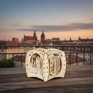 Kerzenhalter aus naturfarbenem Holz für ein Teelicht mit Motiv „Stadt Leipzig“, auf einem Holztisch im Freien platziert. Der filigrane Halter zeigt typische Leipziger Bauwerke und Schriftzug „LEIPZIG“, umgeben von kleinen Sternen, die vom warmen Kerzenlicht im Inneren beleuchtet werden. Im Hintergrund ist eine Stadtansicht mit Dächern und Kirchtürmen bei Sonnenuntergang zu sehen.