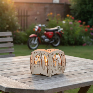 Kerzenhalter aus naturfarbenem Holz für ein Teelicht mit Motiv „Moped“, auf einem Holztisch im Garten platziert. Der filigrane Halter zeigt Ausschnitte mit einem Moped, einer Stadtsilhouette und dekorativen Sternen, die vom warmen Kerzenlicht im Inneren beleuchtet werden. Im Hintergrund steht ein rotes Moped vor Blumen und einem Holzzaun in einer sommerlichen Gartenszene.