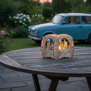 Kerzenhalter aus naturfarbenem Holz für ein Teelicht mit Motiv „Trabant und Wartburg“, auf einem runden Holztisch im Garten platziert. Der filigrane Halter zeigt Ausschnitte mit einer Stadtszene und einem Trabant‑Auto, die vom warmen Kerzenlicht im Inneren beleuchtet werden. Im Hintergrund steht ein hellblauer Trabant vor blühenden Pflanzen und Büschen, eingebettet in eine sommerliche Gartenszene.