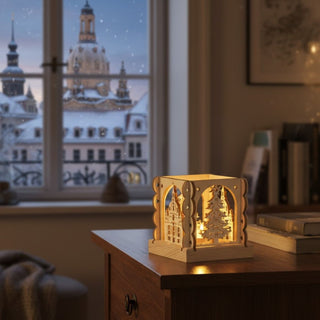 Kleiner Kerzenhalter aus naturfarbenem Holz für ein Teelicht mit Motiv „Dresden“, auf einer Kommode neben Büchern platziert. Filigrane Ausschnitte zeigen die Dresdner Frauenkirche und einen Weihnachtsbaum, die vom warmen Kerzenlicht im Inneren beleuchtet werden; im Hintergrund ist durch ein Fenster die verschneite Dresdner Altstadt bei Abenddämmerung zu sehen.