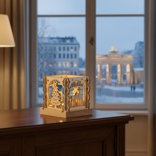 Kleiner Kerzenhalter aus naturfarbenem Holz für ein Teelicht mit Motiv „Berlin“, auf einer Kommode vor einem Fenster platziert. Filigrane Ausschnitte zeigen das Brandenburger Tor, einen Weihnachtsbaum und den Schriftzug „BERLIN“, die vom warmen Kerzenlicht im Inneren beleuchtet werden; im Hintergrund ist eine winterliche Stadtszene zu sehen.