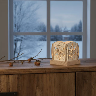 Kleiner Kerzenhalter aus naturfarbenem Holz für ein Teelicht mit Motiv „Weihnachtsmann auf Schlitten“, auf einer Holzkommode platziert. Filigrane Ausschnitte zeigen den Weihnachtsmann mit Rentieren sowie dekorative Sterne, die vom warmen Kerzenlicht im Inneren beleuchtet werden; im Hintergrund ist ein Fenster mit verschneiter Winterlandschaft zu sehen, daneben liegen Zweige mit kleinen Früchten.