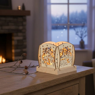 Kleiner Kerzenhalter aus naturfarbenem Holz für ein Teelicht mit Glockenmotiv, auf einer Holzkommode vor einem Fenster platziert. Filigrane Ausschnitte zeigen zwei Glocken mit Schleife und Sternen, die vom warmen Kerzenlicht sanft beleuchtet werden; im Hintergrund sind ein Sessel, ein Kamin und eine verschneite Winterlandschaft zu sehen.