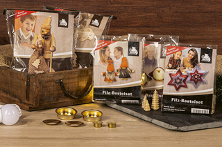 Mehrere Filz‑Bastelsets stehen auf einem Tisch, teilweise in einer offenen Holzkiste. Die Verpackungen zeigen unterschiedliche Bastelprojekte wie Figuren, Sterne und Dekorationen. Vor den Sets liegen kleine Bastelmaterialien wie goldene Metallhalterungen und Zubehörteile.