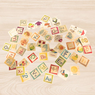 Viele kleine, quadratische Holzplättchen eines farbenfrohen ABC‑Memospiels liegen verstreut auf einem hellen Holztisch; die Plättchen zeigen bunte Buchstabenpaare (z. B. Aa, Bb, Cc) sowie passende Illustrationen und Wörter wie „Apfel“, „Delfin“, „Xylophon“, „Traktor“ oder „Sonne“.