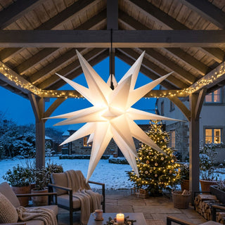 Weißer LED-Adventsstern zum Falten für den Außenbereich von HGD mit 35 cm Durchmesser, aufgehängt unter einer überdachten Terrasse. Der mehrzackige Stern strahlt in warmweißem Licht und setzt einen festlichen Blickfang vor einem winterlichen Garten. Im Hintergrund sorgen ein geschmückter Weihnachtsbaum mit Lichterkanz, verschneite Pflanzen sowie eine gemütliche Sitzgruppe mit Decken und Kerzen für eine stimmungsvolle Advents- und Weihnachtsatmosphäre im Außenbereich.