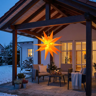 Gelber LED-Adventsstern zum Falten für den Außenbereich von HGD mit 35 cm Durchmesser, aufgehängt unter einer überdachten Terrasse. Der mehrzackige Stern leuchtet in warmem Gelb und setzt einen festlichen Akzent auf der winterlich dekorierten Veranda. Gemütliche Sitzmöbel mit Decken, Kerzen auf dem Beistelltisch und ein leicht verschneiter Garten schaffen eine stimmungsvolle Atmosphäre. Die warme Beleuchtung des Adventssterns spiegelt sich in den Fenstern des Hauses wider.
