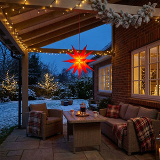 LED-Adventsstern zum Falten für den Außenbereich von HGD in Rot mit 35 cm Durchmesser, aufgehängt unter einer überdachten Terrasse. Der mehrzackige Stern leuchtet in warmem Rot und sorgt für eine festliche Weihnachtsstimmung im Außenbereich. Die gemütliche Lounge mit Polyrattan-Möbeln, karierten Decken, stimmungsvollen Lichterketten und winterlich verschneitem Garten schafft ein einladendes Ambiente für die Advents- und Weihnachtszeit.