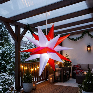 LED-Adventsstern zum Falten für den Außenbereich von HGD in Rot/Weiß mit 35 cm Durchmesser, aufgehängt unter einer überdachten Terrasse. Der dekorative, mehrzackige Stern kombiniert leuchtend rote und weiße Spitzen und strahlt festliches Licht aus. Im Hintergrund sorgen verschneite Tannen, warme Lichterketten, Laternen sowie eine gemütliche Sitzgruppe mit karierten Decken für eine stimmungsvolle Weihnachtsatmosphäre auf der winterlich dekorierten Veranda.