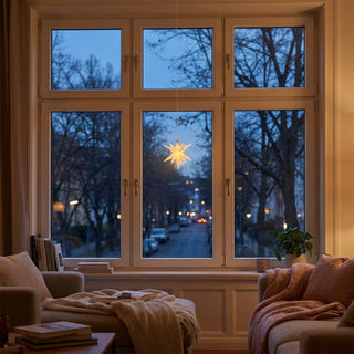 LED Adventsstern von HGD in Weiß mit einem Durchmesser von 8 cm. Der batteriebetriebene Leuchtstern hängt an einer feinen Schnur vor einem großen Fenster und strahlt ein warmweißes Licht aus. Durch die Fensterscheiben ist eine ruhige Straße mit Bäumen und vereinzelten Lichtern in der Dämmerung zu sehen. Der Wohnraum ist gemütlich eingerichtet und verfügt über ein Sofa, einen Sessel, weiche Decken, Bücher und eine Zimmerpflanze.