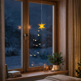 LED Adventsstern von HGD in Gelb mit einem Durchmesser von 8 cm. Der batteriebetriebene Leuchtstern hängt an einer feinen Schnur vor einem großen Holzfenster und strahlt ein warmes, gelbes Licht aus. Auf der Fensterbank stehen Bücher, eine kleine Rentierfigur, ein Teelicht sowie ein dekoratives Tannengesteck mit Zapfen. Im Hintergrund sind eine winterliche Abendlandschaft und die Lichter eines Weihnachtsbaums zu sehen.