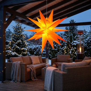 Gelber LED-Adventsstern für Außen von HGD mit 60 cm Durchmesser, aufgehängt unter einer überdachten Terrasse. Der große, mehrzackige Stern strahlt in warmem Gelb und sorgt für eine festliche Weihnachtsstimmung. Im Hintergrund sind verschneite Bäume mit Lichterketten, eine gemütliche Lounge mit Sitzmöbeln, Decken und ein Terrassenheizer zu sehen. Die winterliche Außendekoration schafft eine einladende und stimmungsvolle Atmosphäre zur Adventszeit.