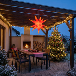 LED-Adventsstern für den Außenbereich von HGD in Rot/Gelb mit 60 cm Durchmesser, aufgehängt unter einer überdachten Terrasse. Der mehrzackige Weihnachtsstern leuchtet in warmen Rot- und Gelbtönen und sorgt für festliche Stimmung. Neben einem beleuchteten Weihnachtsbaum mit roten Kugeln sind eine gemütliche Sitzgruppe, ein Kaminfeuer und stimmungsvolle Lichterketten zu sehen. Die winterlich dekorierte Terrasse schafft eine einladende Atmosphäre für die Advents- und Weihnachtszeit.