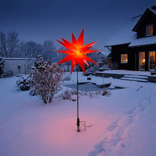 LED Adventsstern von HGD in leuchtendem Rot mit einem Durchmesser von 60 cm. Der beleuchtete Stern ist auf einem schwarzen Metallstab mit Bodenspieß montiert und steht in einem verschneiten Garten vor einem Haus in der Dämmerung. Das warme Licht des Sterns setzt einen festlichen Akzent und hebt sich deutlich von der winterlichen, blau getönten Umgebung ab. Der Bodenspieß ermöglicht eine einfache Platzierung im Außenbereich.