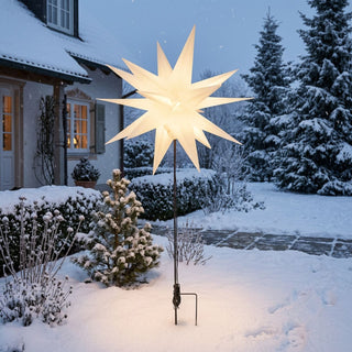 LED Adventsstern für den Außenbereich von HGD in Weiß mit einem Durchmesser von 60 cm. Der beleuchtete Weihnachtsstern ist auf einem schwarzen Metallstab mit Bodenspieß montiert und in einem verschneiten Vorgarten vor einem Wohnhaus aufgestellt. Das warmweiße Licht des Sterns sorgt für eine festliche Atmosphäre und bildet einen schönen Kontrast zur winterlichen Umgebung mit schneebedeckten Sträuchern, Nadelbäumen und Wegen. Der Bodenspieß ermöglicht eine stabile und flexible Platzierung.