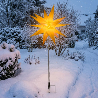 LED Adventsstern für den Außenbereich von HGD in leuchtendem Gelb mit 60 cm Durchmesser. Der beleuchtete Weihnachtsstern ist an einem schlanken Metallstab mit Bodenspieß befestigt und steht in einem verschneiten Garten. Das warme gelbe Licht des Sterns kontrastiert mit der winterlichen Umgebung aus schneebedeckten Sträuchern, Bäumen und Wegen. Die Dekoration eignet sich zur stimmungsvollen Beleuchtung von Garten, Vorgarten oder Eingangsbereich in der Advents- und Weihnachtszeit.