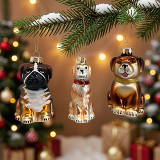 Drei Hunde aus Glas als Christbaumschmuck hängen an einem verschneiten Tannenzweig vor festlich beleuchteter Weihnachtskulisse.