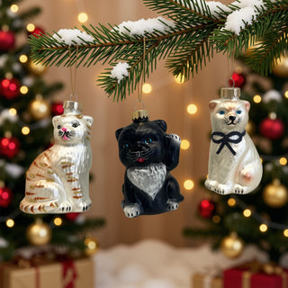 Drei Katzen aus Glas als Christbaumschmuck hängen an einem verschneiten Tannenzweig vor festlich beleuchtetem Weihnachtsdekor.