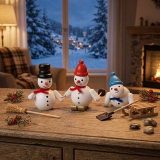 Miniatur „Schneemänner“, 3er Set (je ca. 6 cm), farbig gestaltet mit drei detailreichen Figuren, jeweils mit Mütze und Schal, teilweise mit Besen, Schaufel und Schlitten. Die Schneemänner stehen auf einem Holztisch in einer gemütlichen Wohnstube; im Hintergrund sind ein Fenster mit verschneiter Winterlandschaft und ein Kamin zu sehen, ergänzt durch winterliche Deko wie Zweige und Beeren.