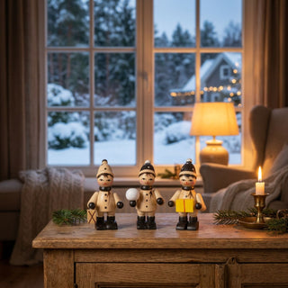 Miniatur „Winterkinder“, 3er Set (je ca. 6 cm), aus naturbelassenem Holz mit drei Figuren in Winterkleidung, jeweils mit kleinen Accessoires wie Laterne, Schneeball und Geschenk. Die Figuren stehen auf einem Holztisch in einer gemütlichen Stube; im Hintergrund ein Fenster mit verschneiter Winterlandschaft, eine Lampe und eine Kerze sorgen für eine warme, stimmungsvolle Atmosphäre.