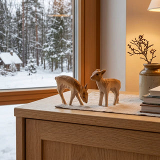 Miniatur „Rehe“, 2er Set (ca. 4 cm), naturbelassen und filigran geschnitzt, mit zwei kleinen Rehfiguren in unterschiedlichen Positionen. Die Figuren stehen auf einem Holzmöbel neben einem Fenster mit verschneiter Winterlandschaft; eine Vase mit Zweigen und eine warme Lampe schaffen eine ruhige, winterliche Atmosphäre.