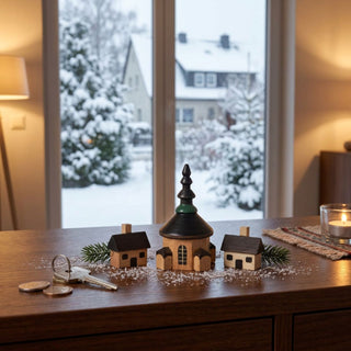 Miniatur „Seiffener Kirche mit 2 Häusern“, 3-teilig (ca. 5 cm), aus naturbelassenem Holz mit einer detailreichen Kirche und zwei kleinen Häusern. Die Szenerie steht auf einem Holzmöbel vor einem Fenster mit verschneiter Winterlandschaft; Tannenzweige, eine Kerze sowie kleine Alltagsgegenstände sorgen für eine ruhige, gemütliche Atmosphäre.